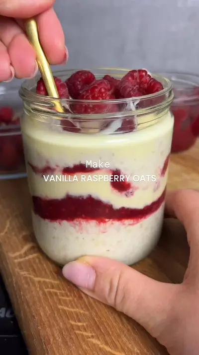 Eine Hand hält ein Glas mit geschichteten Overnight Oats, garniert mit frischen Himbeeren und einem goldenen Löffel.