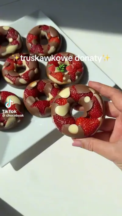 Eine Hand hält einen mit Erdbeeren und Schokolade verzierten Donut über einem Teller mit weiteren Donuts.
