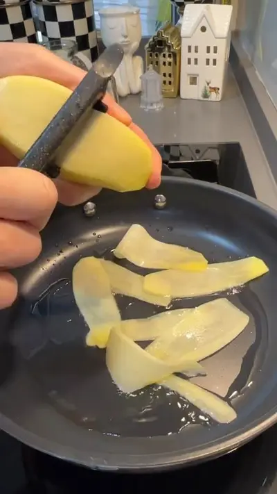 Eine Hand schneidet mit einem Sparschäler dünne Kartoffelscheiben direkt in eine heiße Pfanne mit Öl.