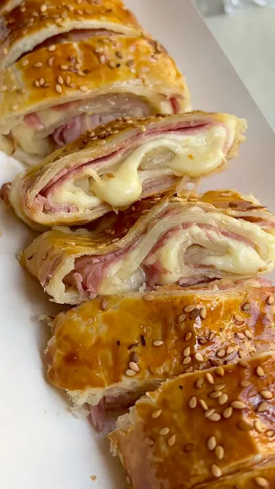 Tranches dorées d'un roulé feuilleté au jambon et fromage fondu parsemé de graines de sésame sur fond blanc.