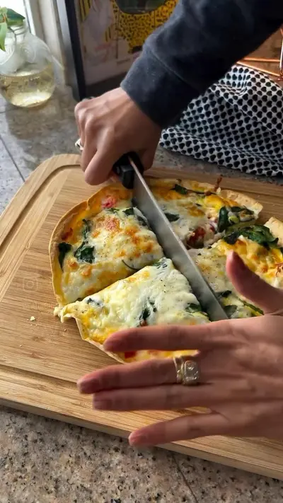 Eine Person schneidet eine goldbraun gebackene Tortilla-Quiche mit Käse, Spinat und Tomaten auf einem Holzbrett.