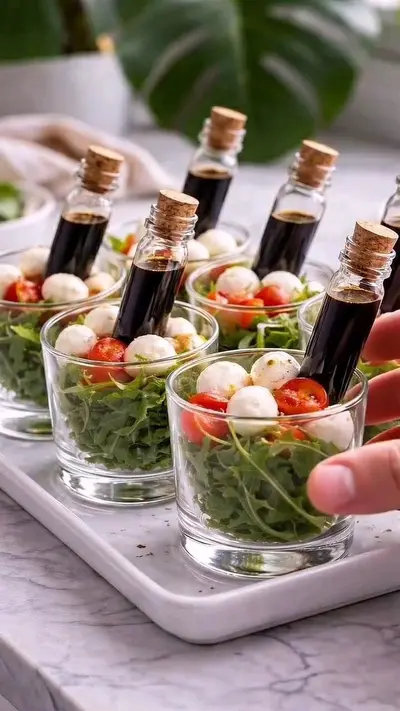 A close-up of small glass cups filled with arugula, cherry tomatoes, and mozzarella pearls, each holding a balsamic vial.