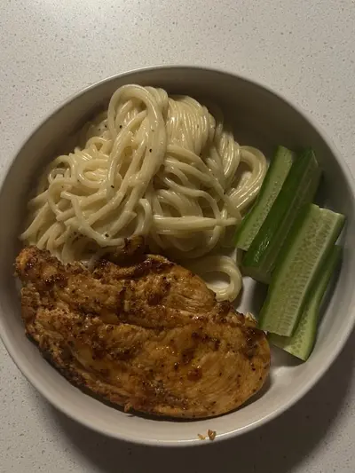Ein Teller mit gebratenem Hähnchen, Spaghetti in cremiger Soße und frischen Gurkenstücken.
