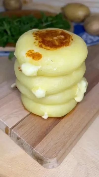 Ein Stapel von vier goldbraun gebratenen Kartoffelpuffern mit schmelzendem Käsekern auf einem Holzbrett.