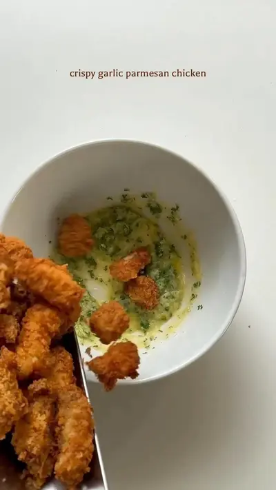 A metal spatula dropping golden fried chicken pieces into a bowl filled with a garlic herb and parmesan butter sauce.