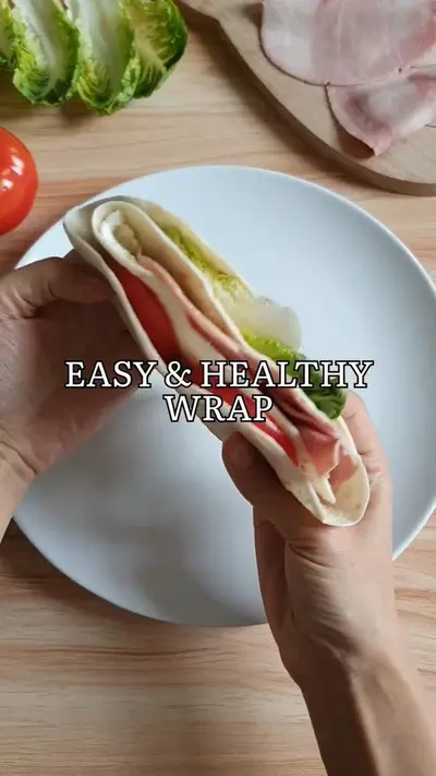 Mains tenant un wrap frais garni de jambon, laitue et tomate au-dessus d'une assiette blanche.