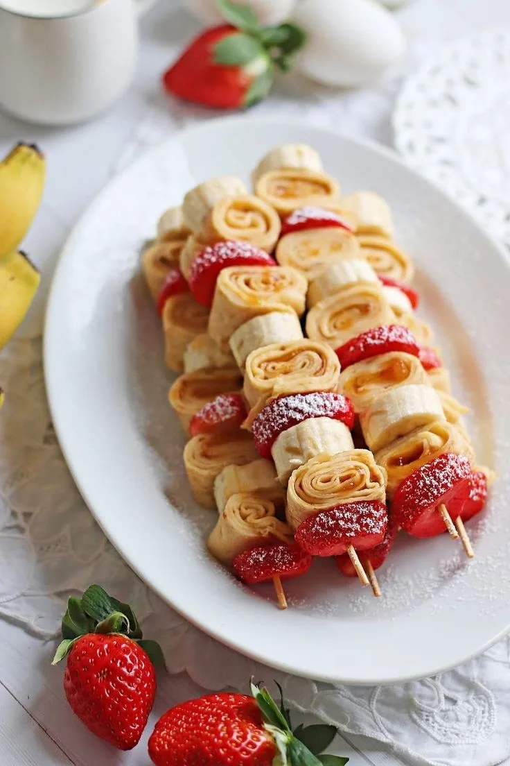 Ein weißer Teller mit Crepe-Rollen, Bananen und Erdbeeren auf Holzspießen, bestäubt mit Puderzucker.