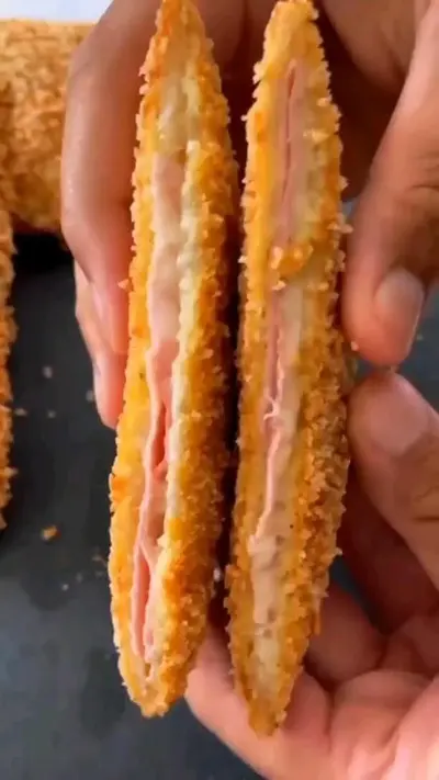 Twee handen houden een doorgesneden, gefrituurde snack vast met een krokante buitenkant en gesmolten kaas en ham.